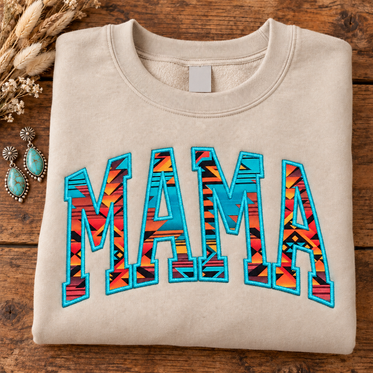 Sand Western MAMA Aztec appliqué crewneck folded on a rustic wood table with turquoise western earrings; multicolor Aztec fabric letters with turquoise embroidery