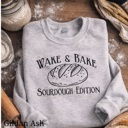 Gray sweatshirt with 'Wake & Bake Sourdough Edition' text and bread graphic on a wooden surface with baking ingredients.