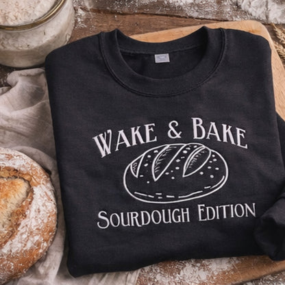 Black sweatshirt with 'Wake & Bake Sourdough Edition' design on a wooden surface with bread and flour.