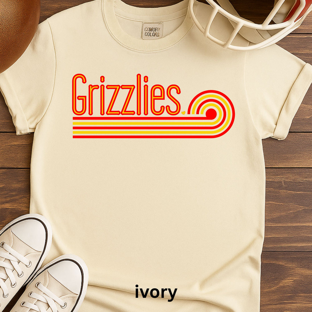 An ivory short-sleeve t-shirt with a retro-style 'Grizzlies' graphic in red and yellow, styled in a flat-lay on a wooden surface with a football helmet, sneakers, and a leather ball partially visible.