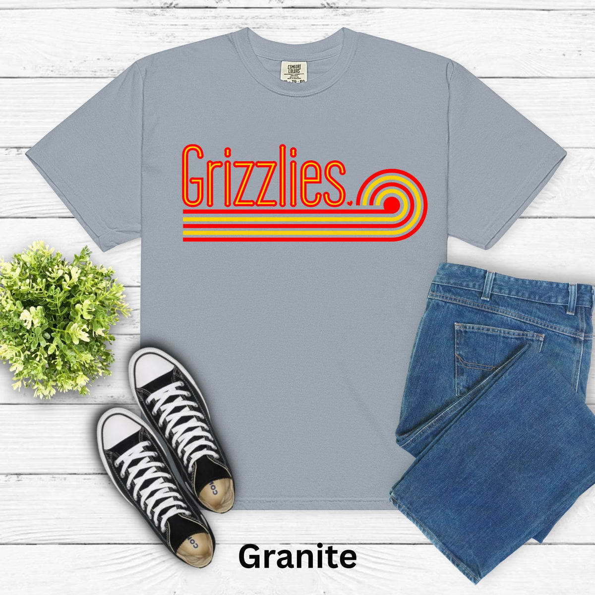 A flat-lay of a light gray short-sleeve t-shirt with a retro-style 'Grizzlies' graphic in red and yellow. The shirt is styled with blue jeans, black and white sneakers, and a small green plant on a white wood plank background.
