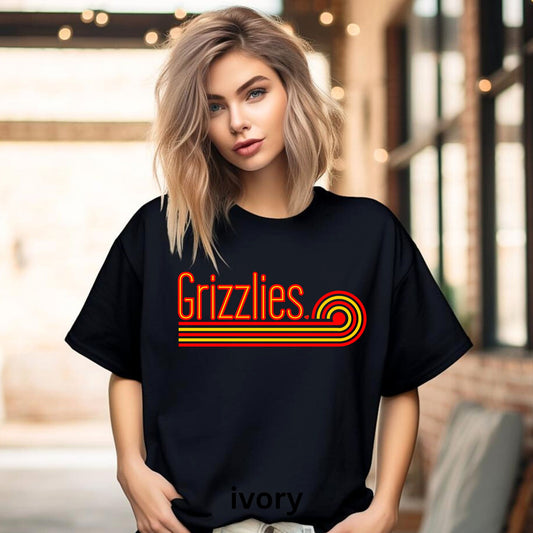 A woman wearing a black oversized T-shirt with a retro-style red and orange 'Grizzlies' graphic across the chest, standing in a softly blurred indoor setting with warm lights and brick walls.