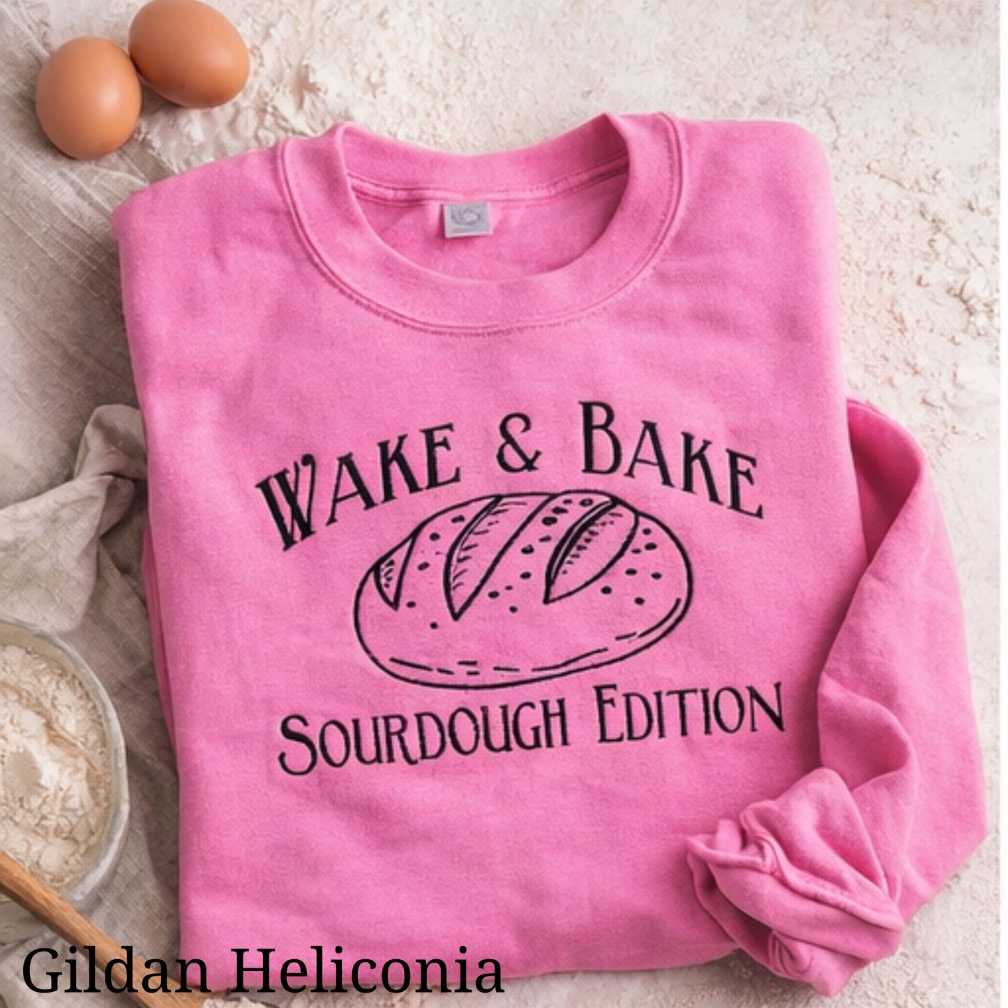 Pink sweatshirt with 'Wake & Bake Sourdough Edition' text and bread graphic on a flour-dusted surface.