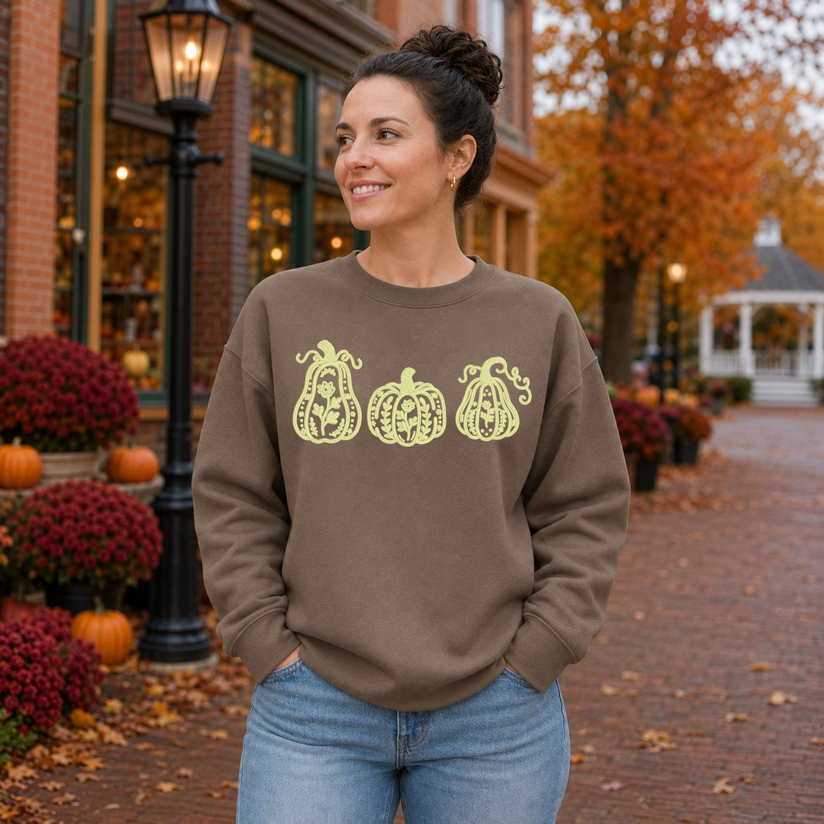 Woman wearing a Brown Savana Gildan Softstyle sweatshirt embroidered with a cream botanical pumpkin trio in a New England fall town square