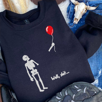 Black Gildan sweatshirt with skeleton and red balloon embroidery styled in a western flat lay with denim accessories.