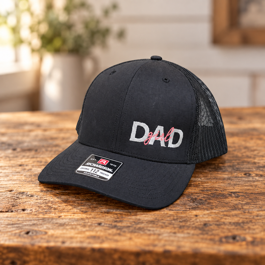 Black Richardson 112 trucker hat with white and pink Girl Dad embroidery on a rustic wood table