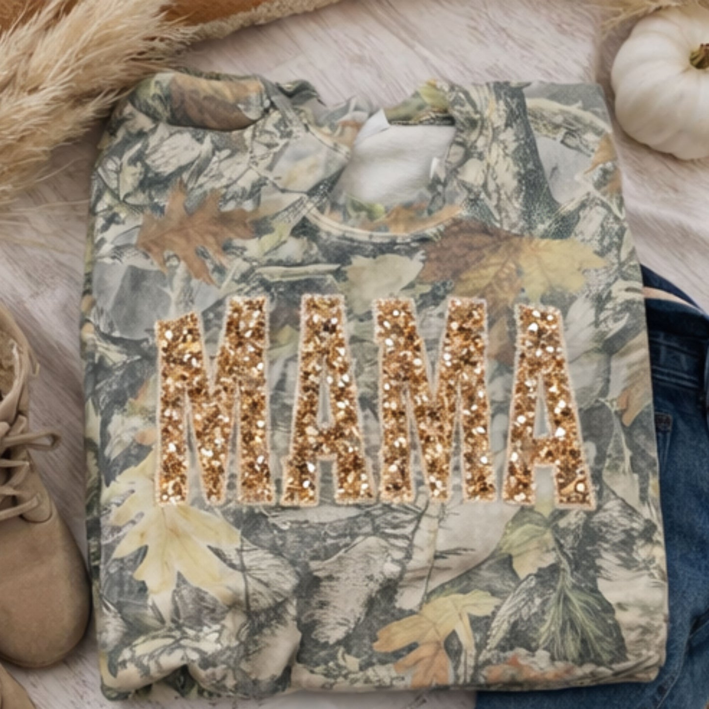 Camouflagecrewneck with 'MAMA' in sequins on a wooden surface with pumpkins and leaves.