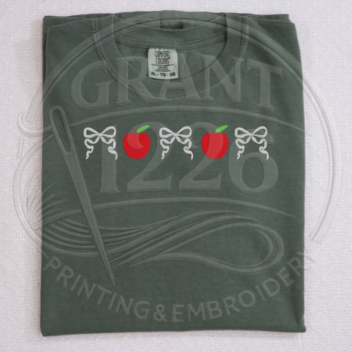 Folded moss green teacher shirt with embroidered apple and bow design