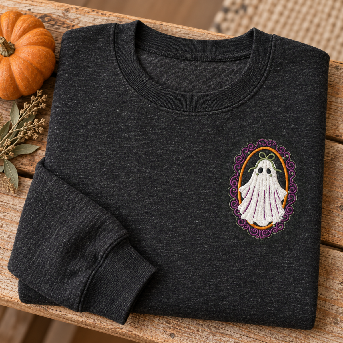 Folded heathered-black ghost sweatshirt on a rustic bench with a pumpkin and dried sage.
