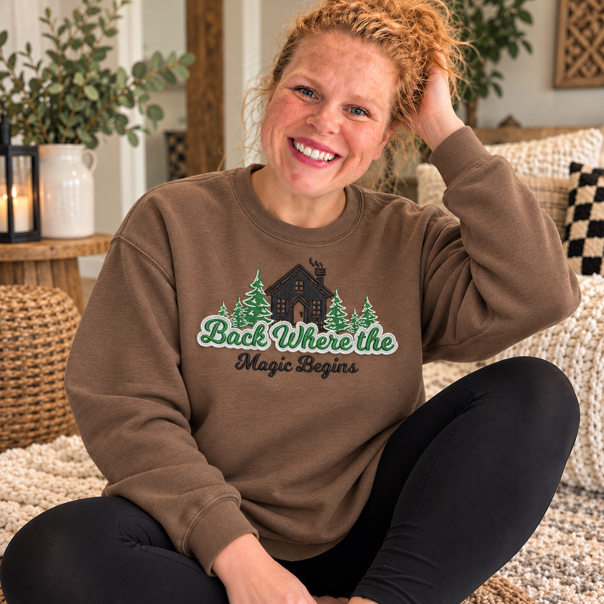 Brown Savanna Gildan Softstyle crewneck sweatshirt with a full-chest embroidered cabin, evergreen trees, and Back Where the Magic Begins design, worn by Samantha indoors.