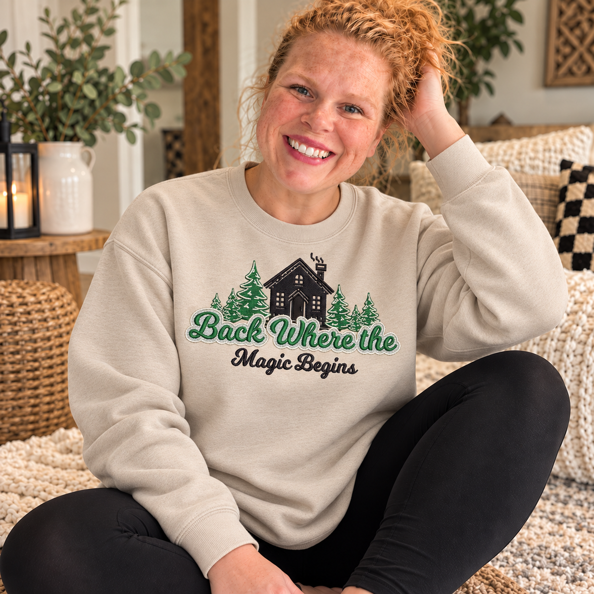 Sand Gildan Softstyle crewneck sweatshirt with a full-chest embroidered cabin, evergreen trees, and Back Where the Magic Begins design, worn by Samantha indoors.