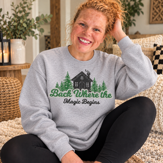 Sport Grey Gildan Softstyle crewneck sweatshirt with a full-chest embroidered cabin, evergreen trees, and Back Where the Magic Begins design, worn by Samantha indoors.