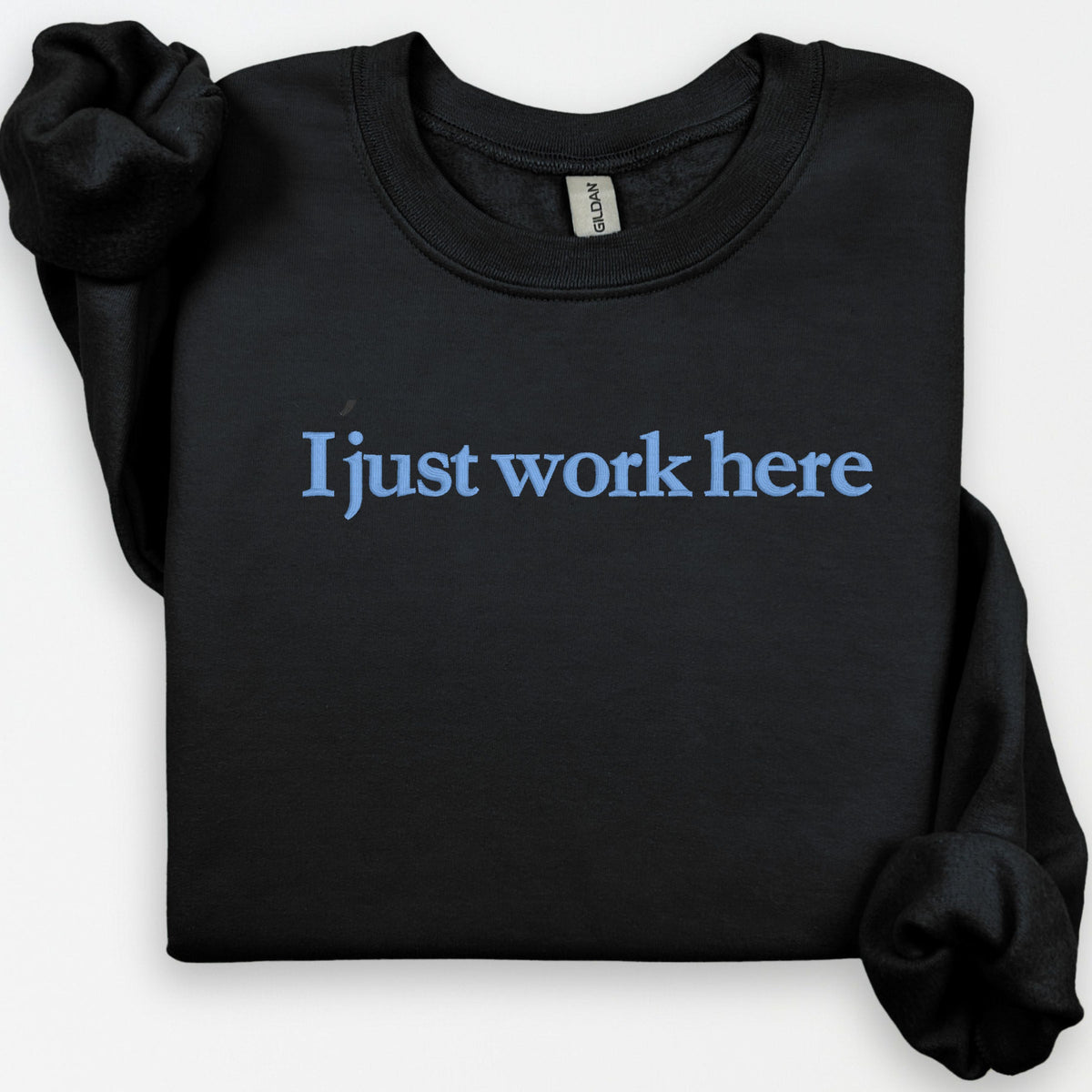 Black Gildan crewneck sweatshirt embroidered with I Just Work Here in a flat-lay product view