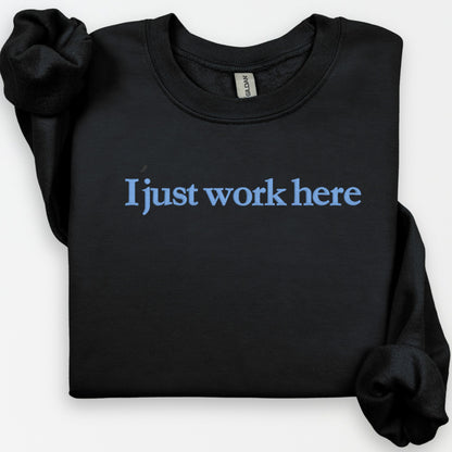 Black Gildan crewneck sweatshirt embroidered with I Just Work Here in a flat-lay product view
