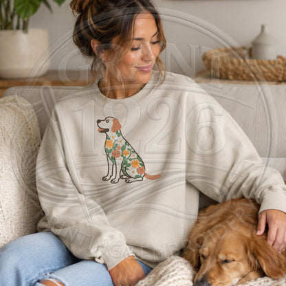 Woman wearing an ivory floral Labrador Retriever sweatshirt while sitting at home beside a dog.