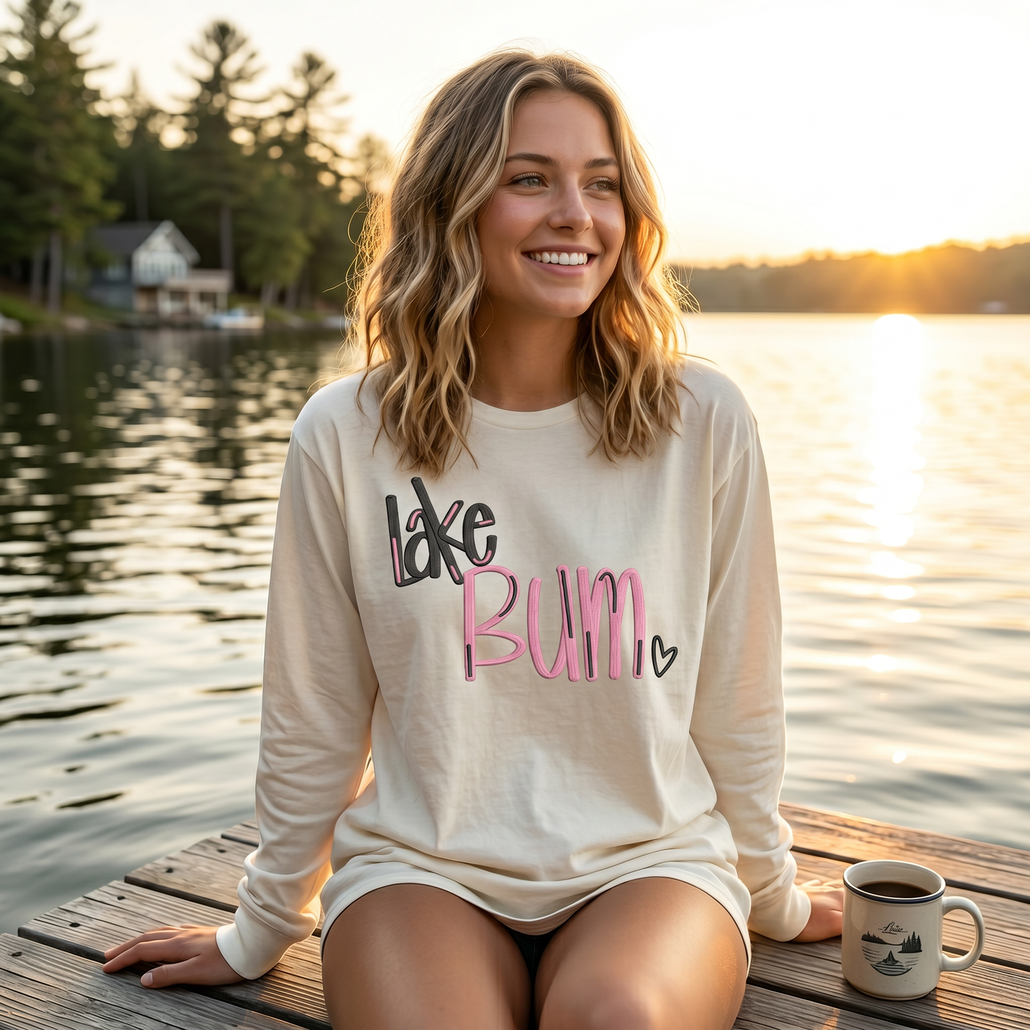 Woman wearing cream embroidered Lake Bum long sleeve shirt sitting on dock by lake