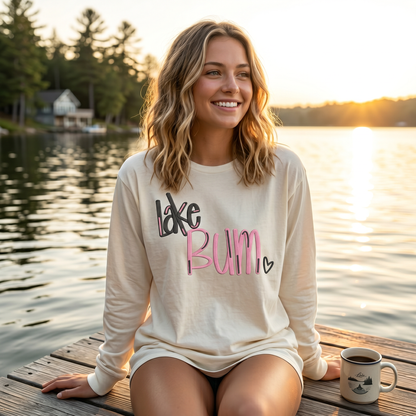 Woman wearing cream embroidered Lake Bum long sleeve shirt sitting on dock by lake