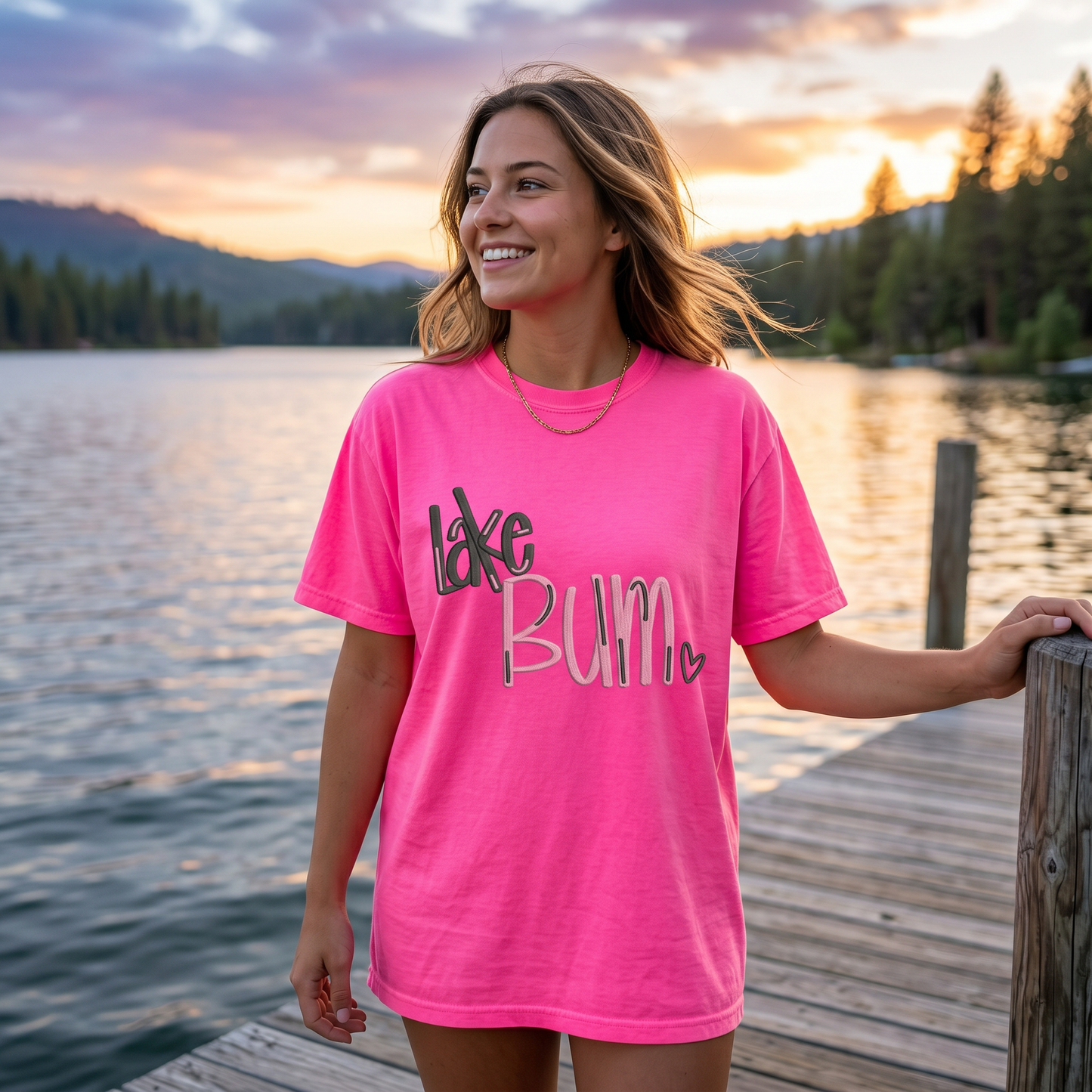Woman wearing bright pink embroidered Lake Bum shirt standing on dock at sunset by the lake