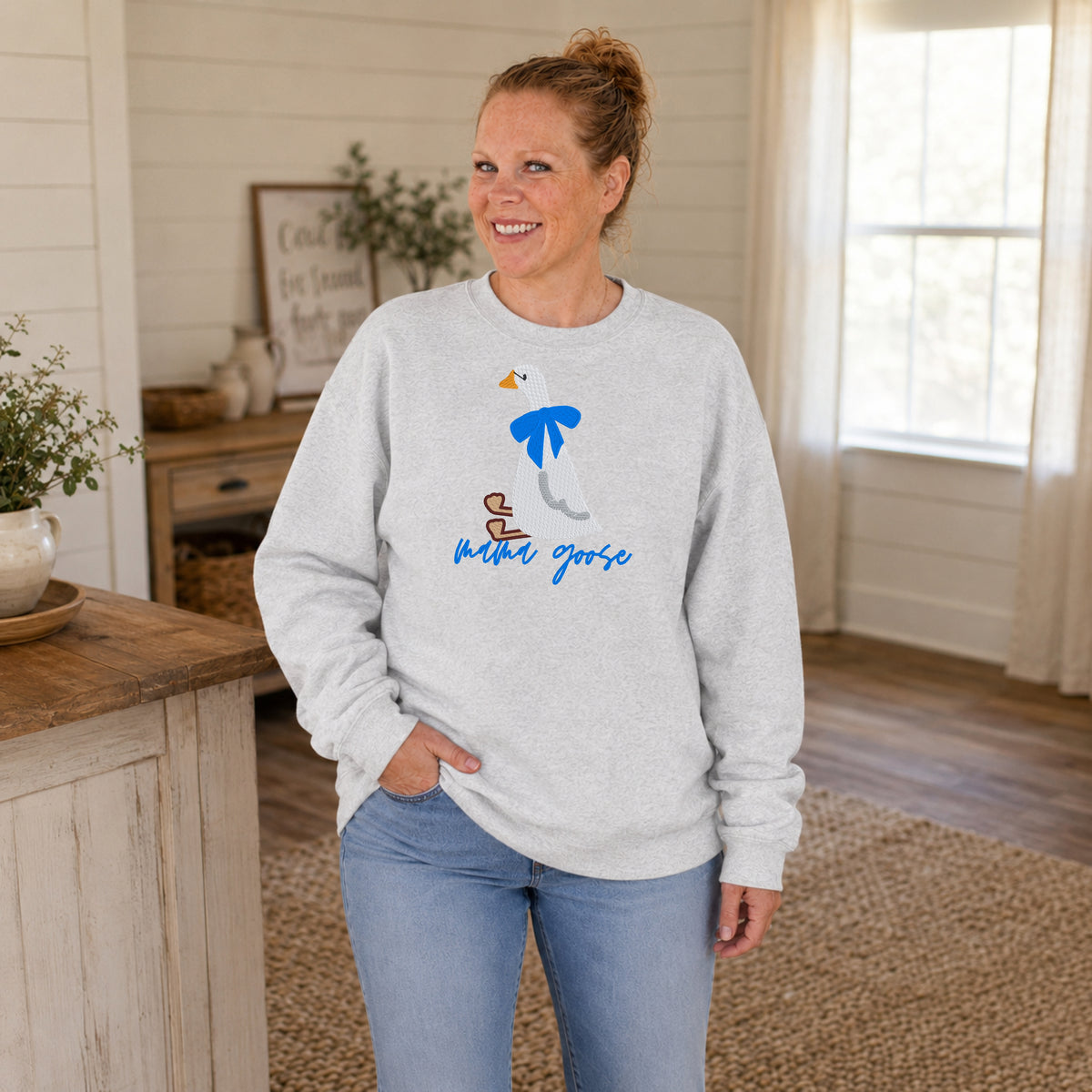 Woman wearing an ash gray Mama Goose crewneck featuring an embroidered white goose with a blue bow.