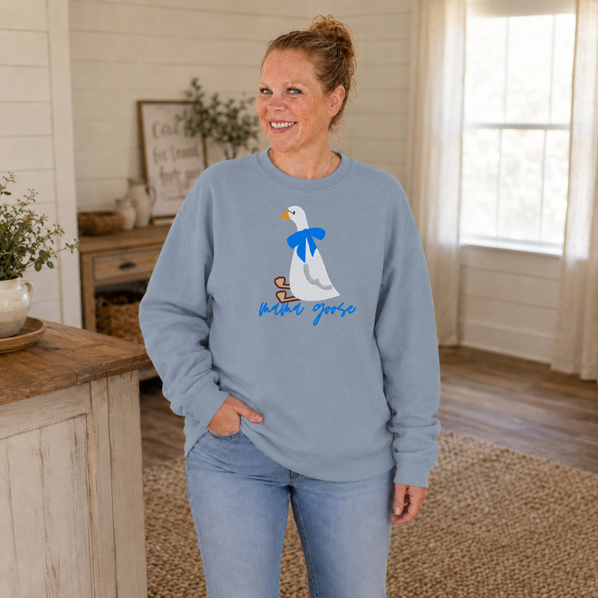 Woman wearing a dusty blue crewneck embroidered with a white goose, blue bow, and Mama Goose lettering.