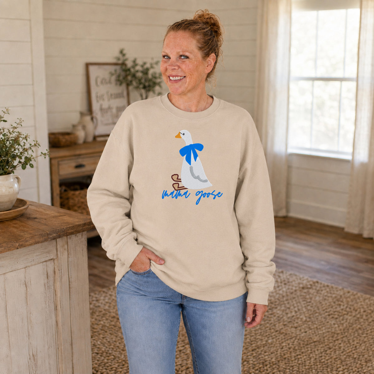Woman modeling a sand-colored Mama Goose sweatshirt with an embroidered goose and bright blue bow.