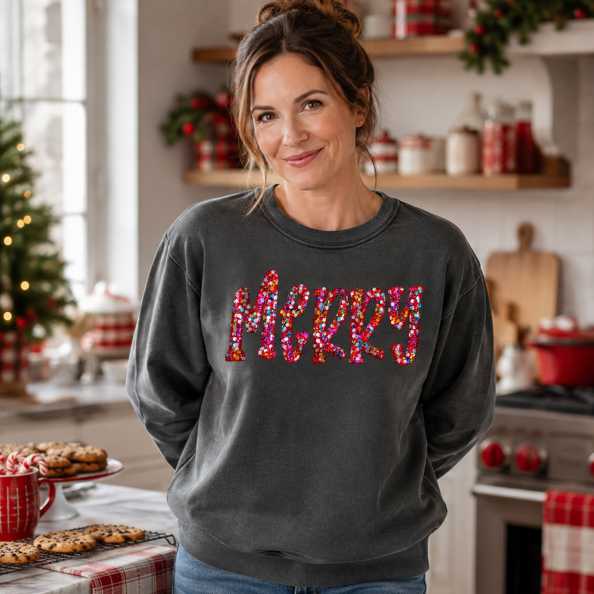 Mockup of a woman wearing a charcoal MERRY sweatshirt with multicolored sequin lettering in a Christmas kitchen