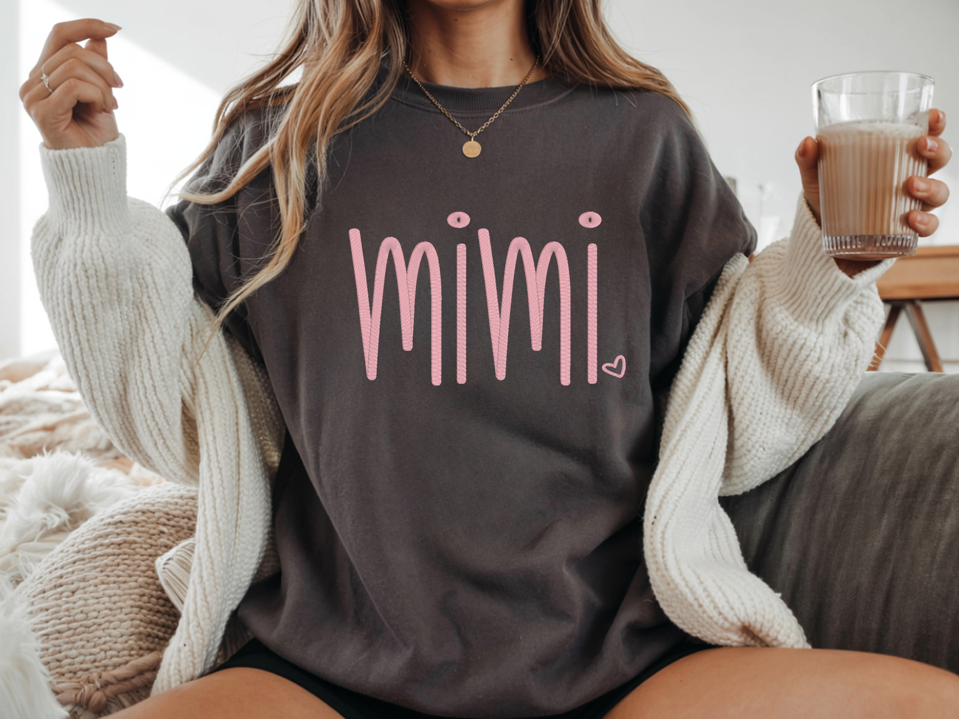 Person wearing a dark gray tshirt with 'mimi' in pink embroidery, holding a glass of coffee.