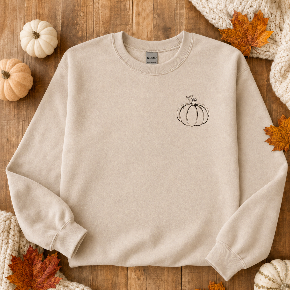 Sand Gildan Softstyle sweatshirt flat lay with small black pumpkin embroidery and autumn leaves