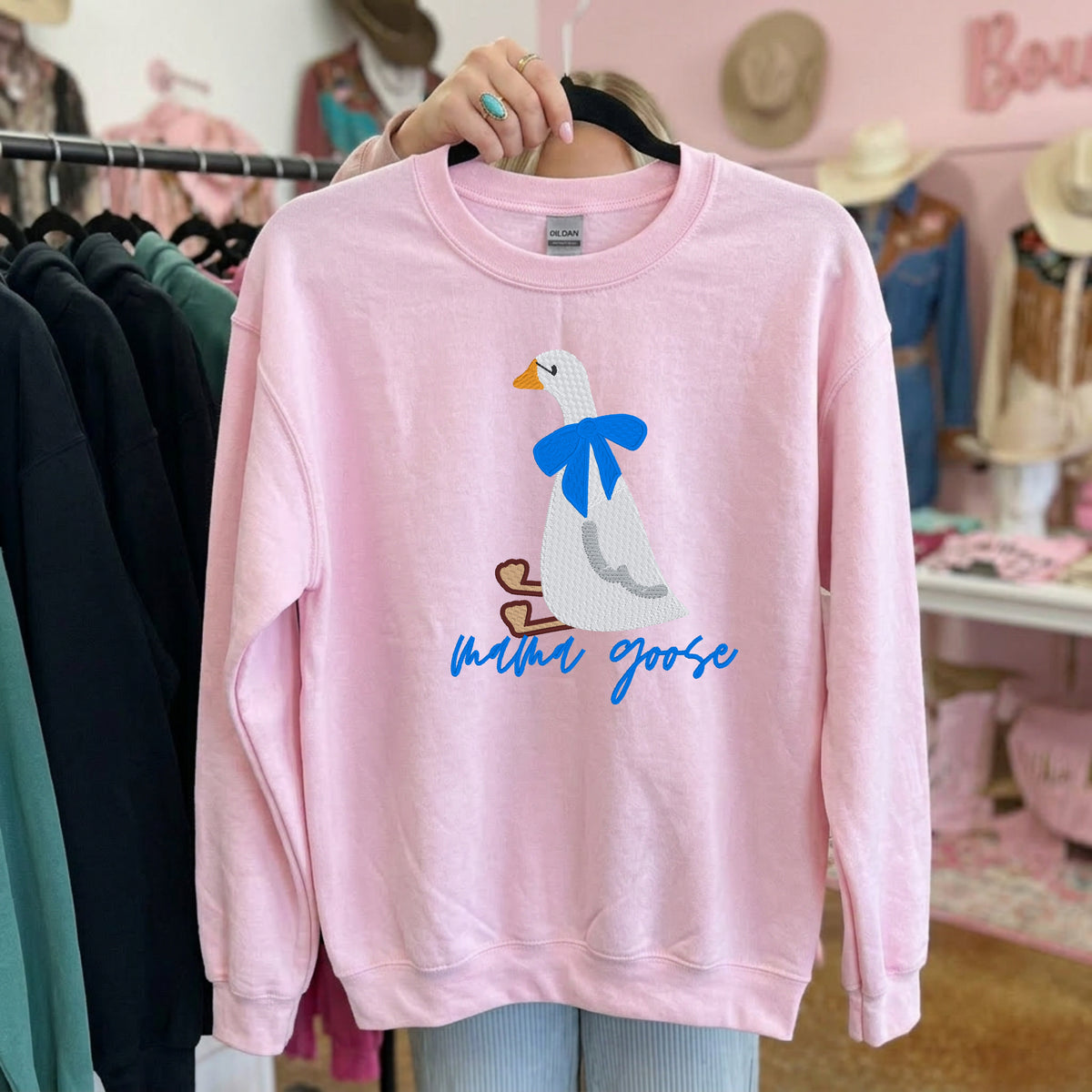Light pink crewneck on a hanger featuring an embroidered white goose, blue bow, and Mama Goose script.