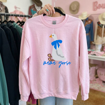 Light pink crewneck on a hanger featuring an embroidered white goose, blue bow, and Mama Goose script.