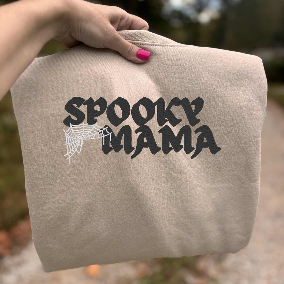 Close view of black Spooky Mama embroidery and white spiderweb stitching on sand crewneck sweatshirt