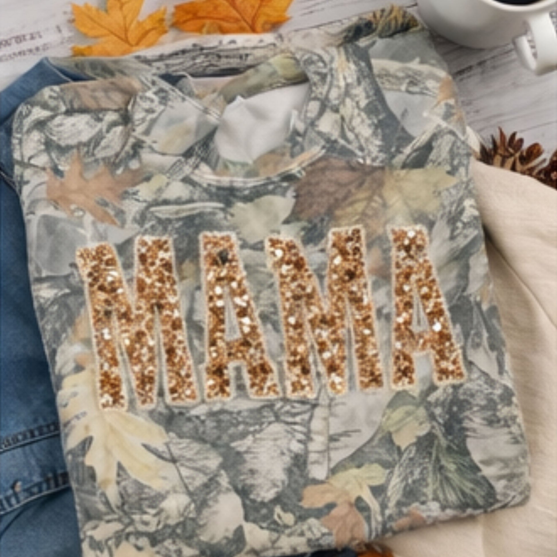 Camouflage sweatshirt with 'MAMA' in sequins on a wooden surface with autumn leaves and a mug.