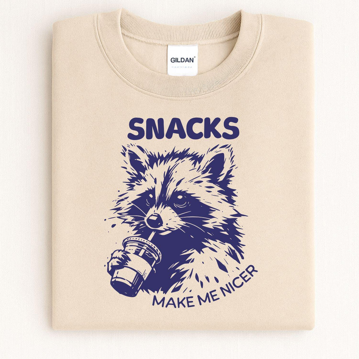 Folded sand crewneck with Snacks Make Me Nicer raccoon graphic