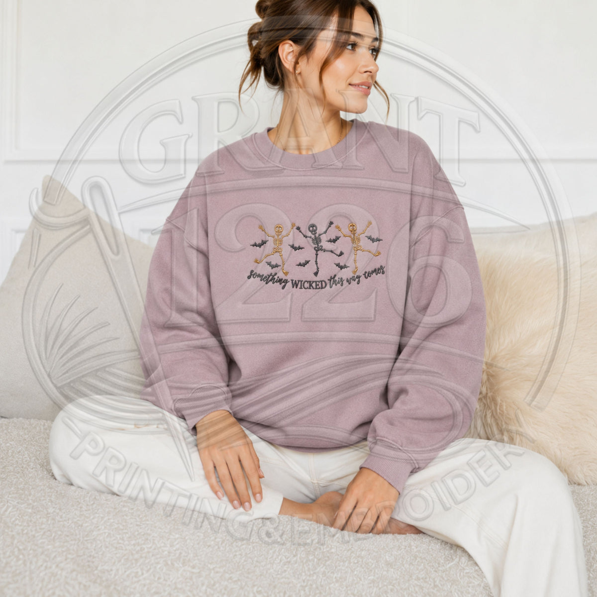 Woman wearing mauve embroidered Halloween skeleton sweatshirt with dancing skeletons, bats and Something Wicked This Way Comes text