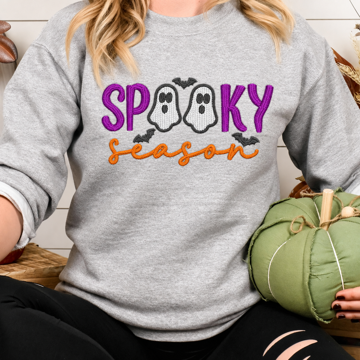Woman wearing a Spooky Season ghost embroidered gray sweatshirt beside a green pumpkin
