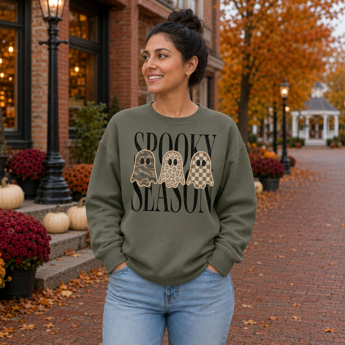 Woman wearing a Military Green Gildan Softstyle Spooky Season patchwork ghost sweatshirt in a fall town