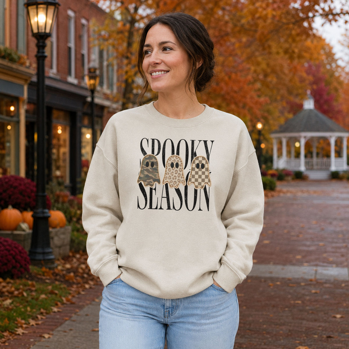 Woman wearing a Sand Gildan Softstyle Spooky Season patchwork ghost sweatshirt in a fall town