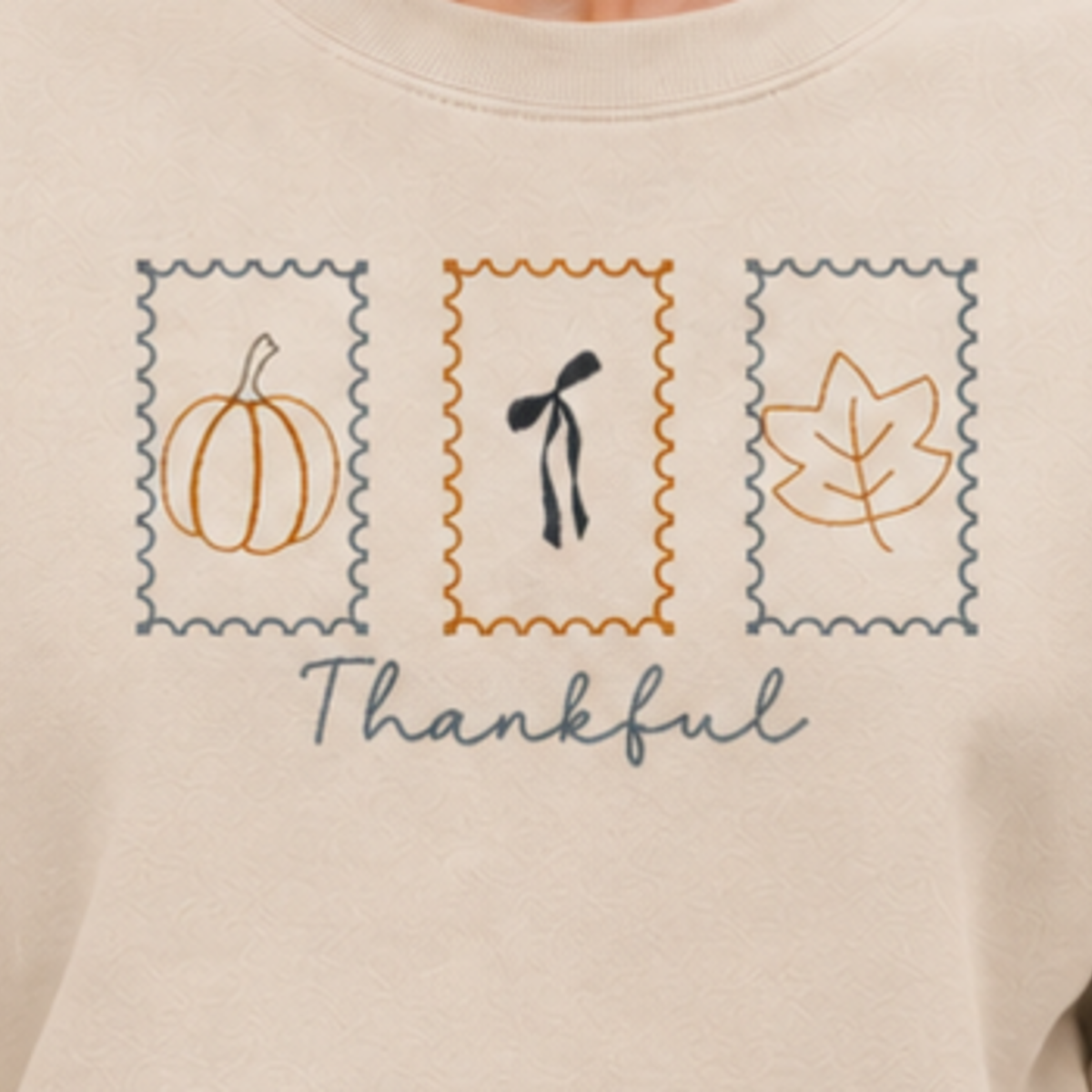 Close-up of Thankful Stamps pumpkin, bow, and leaf embroidery on Sand sweatshirt