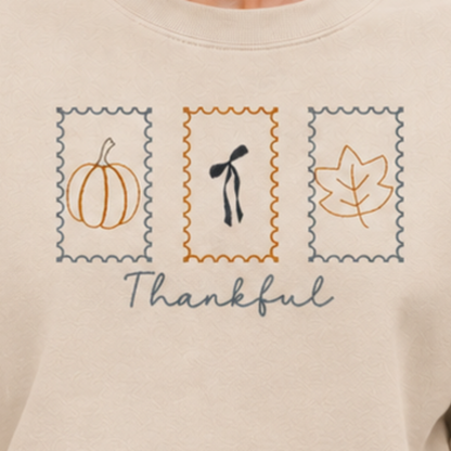 Close-up of Thankful Stamps pumpkin, bow, and leaf embroidery on Sand sweatshirt