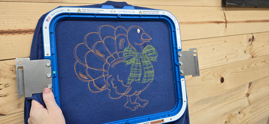 Copper-colored turkey outline with an oversized green plaid bow embroidered on China Blue fabric, shown in an embroidery hoop.