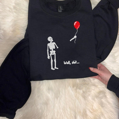 Folded black Gildan crewneck featuring an embroidered skeleton holding a red balloon with “Well, shit…” text.