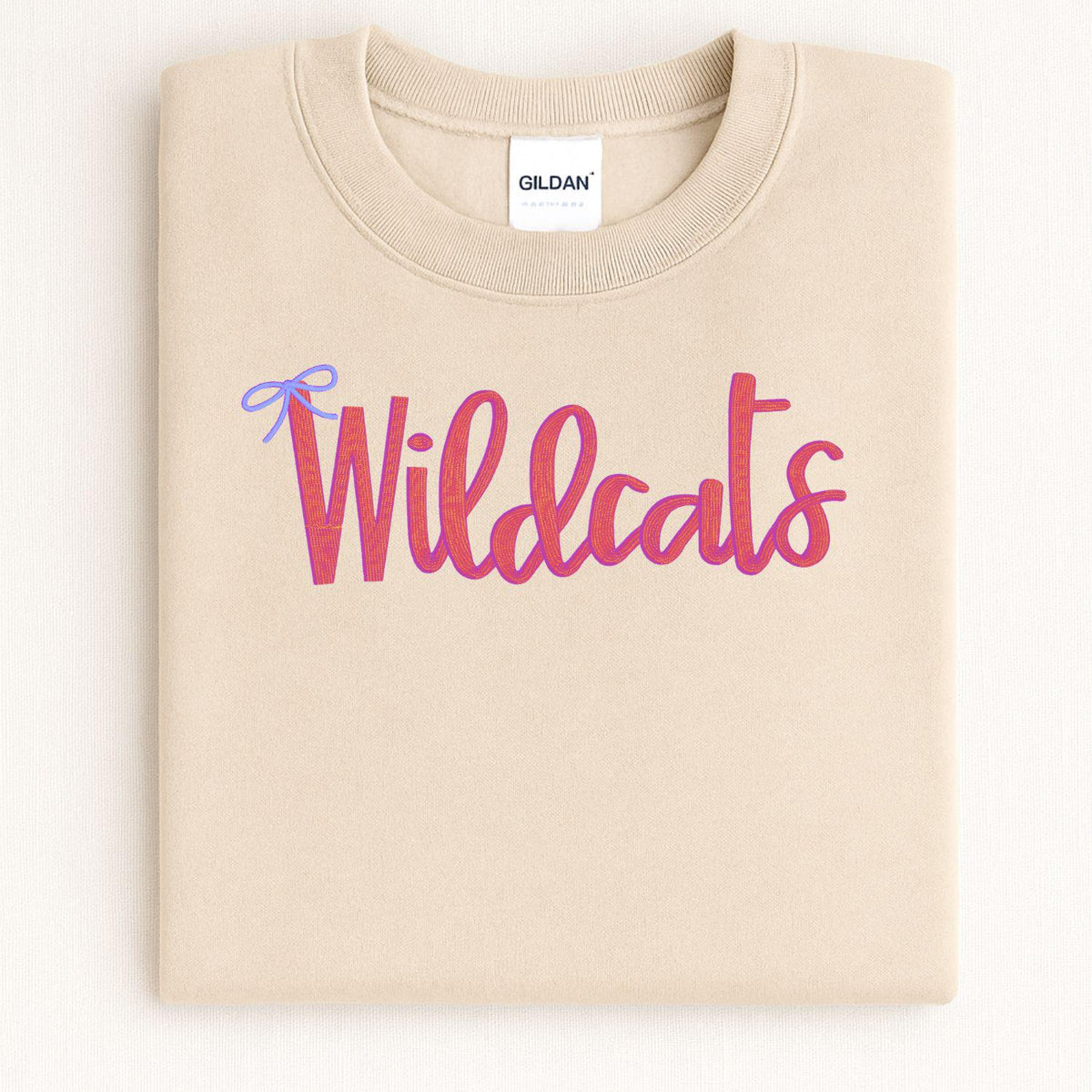 Folded sand crewneck with pink Wildcats script and blue bow embroidery