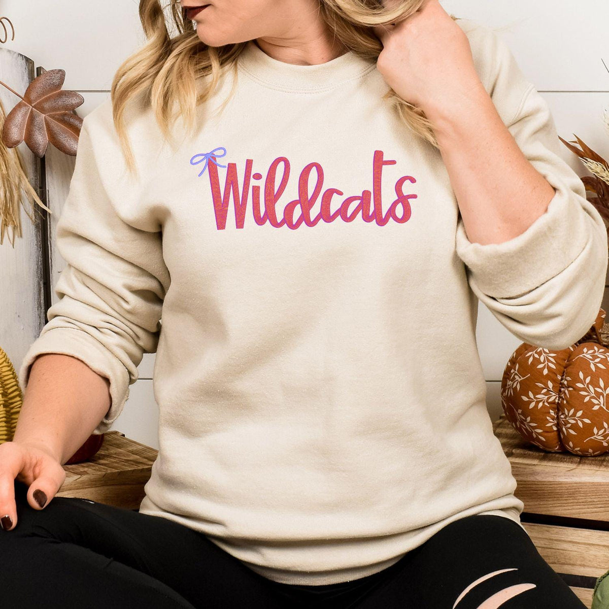Model wearing sand Wildcats script sweatshirt with pink lettering and blue bow