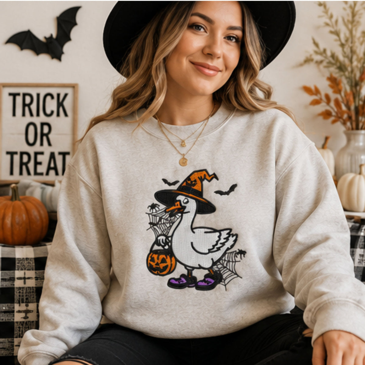 Woman in embroidered witch goose Halloween crewneck with bats, pumpkin trick-or-treat bucket and spiderwebs